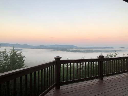 A serene view from a wooden deck overlooking misty mountains at dawn, with a soft pastel sky and a rising moon.