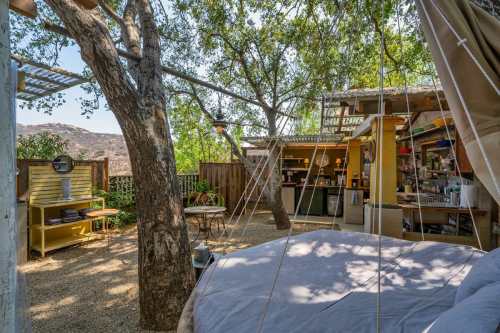 Cozy outdoor space with a bed, tree, and rustic kitchen area surrounded by greenery and mountains.