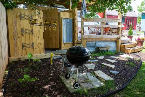 A cozy outdoor space featuring a grill, stone pathway, and wooden fencing surrounded by greenery.
