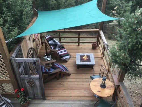 A cozy wooden deck with a green shade sail, seating area, fire pit, and potted plants surrounded by trees.