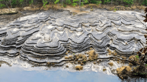 Layered geological formations with wavy patterns, surrounded by sparse vegetation and a small body of water.