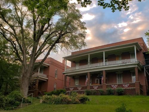 Historic red brick building with balconies, surrounded by greenery and trees, under a cloudy sky.