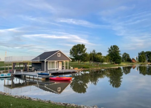 A serene lakeside scene featuring a small gray house, kayaks, and trees reflected in calm water under a blue sky.