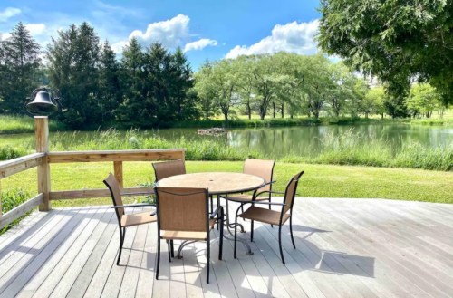 A serene outdoor scene featuring a round table and chairs on a deck, overlooking a pond and lush greenery.