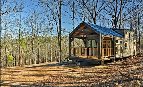 A small wooden cabin with a blue roof, surrounded by bare trees and a clear sky, set in a natural landscape.