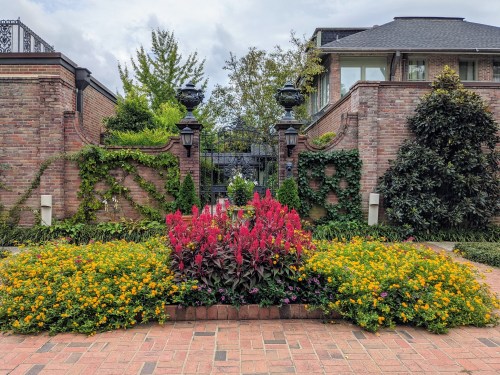 A vibrant garden with colorful flowers in front of a brick wall and ornate gate, surrounded by lush greenery.
