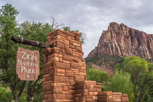 Sign for Zion National Park with a rocky mountain backdrop and lush greenery.