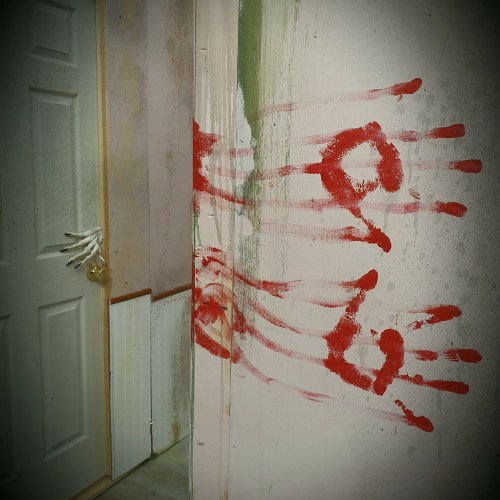 A dimly lit hallway with a door and red handprints smeared on the wall, creating a spooky atmosphere.