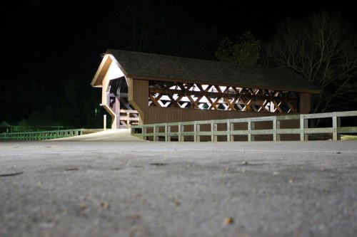 A lit covered bridge at night, surrounded by trees and a pathway leading towards it.