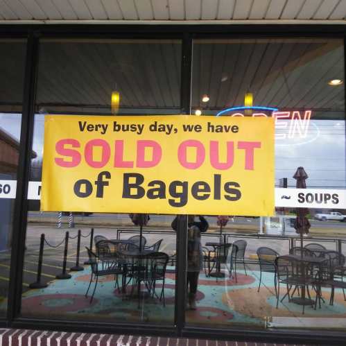 A bright yellow sign in a window reads, "Very busy day, we have SOLD OUT of Bagels." Tables are visible outside.