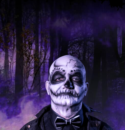 A person with a skull face paint stands in a dark, misty forest, wearing a black coat and bow tie.