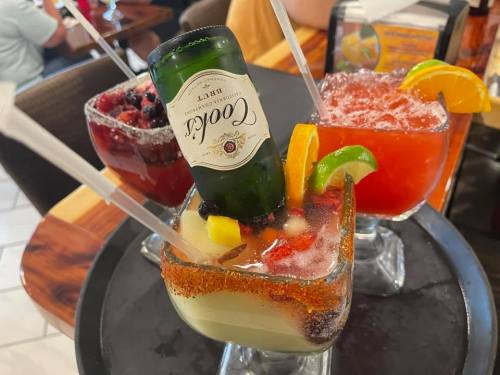 Three colorful cocktails on a tray, each garnished with fruit and one featuring a beer bottle.