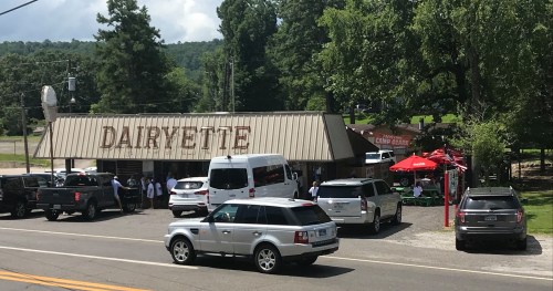 A small roadside diner named "Dairyette" with outdoor seating and several cars parked nearby. Trees in the background.