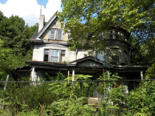 An old, dilapidated house surrounded by overgrown greenery and trees under a partly cloudy sky.