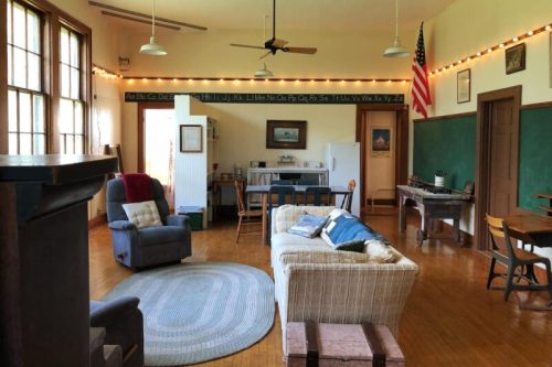 Cozy room with a sofa, chairs, and a kitchen area, featuring chalkboards and an American flag. Warm, inviting atmosphere.