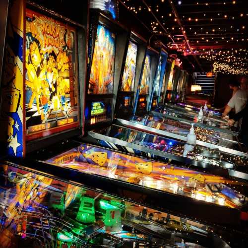 A row of colorful pinball machines illuminated by string lights in a vibrant arcade setting.