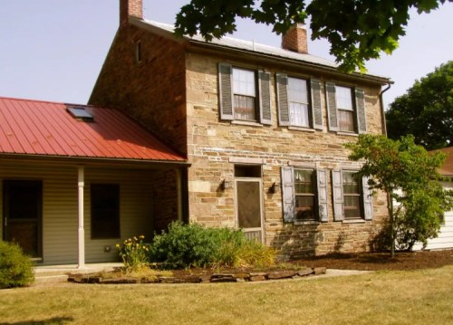 A two-story stone house with a red metal roof, green shutters, and a porch surrounded by landscaping.