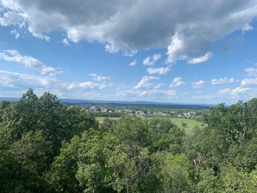 A scenic view of green trees and fields under a blue sky with fluffy clouds, stretching towards distant mountains.