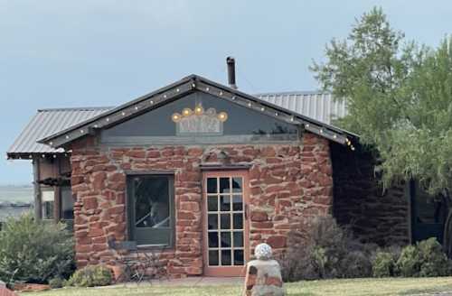 A rustic stone building with a metal roof, adorned with string lights, surrounded by greenery and a small seating area.