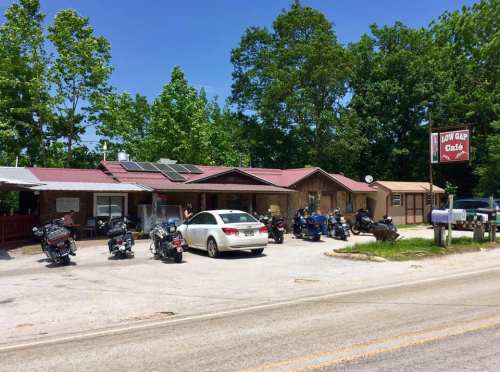 A rustic café with a red sign, surrounded by trees, featuring parked motorcycles and a car in the foreground.
