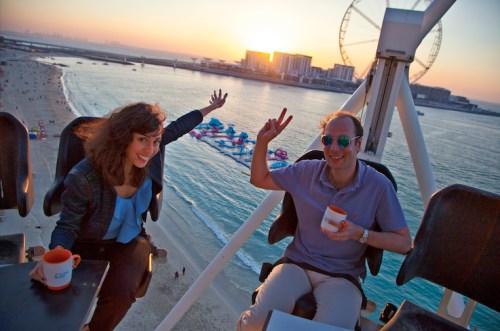 A man and woman enjoy a sunset view from a Ferris wheel, holding cups and smiling, with a beach and city skyline in the background.