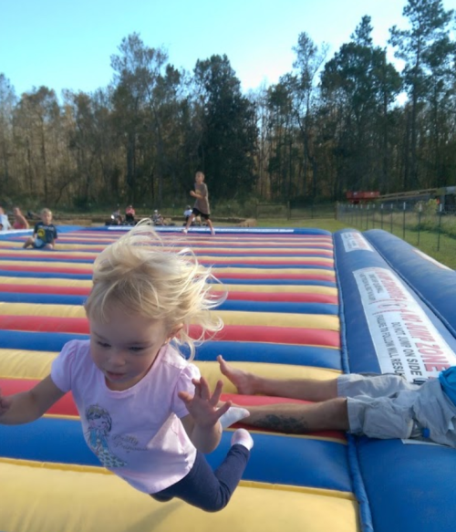 A young child with blonde hair jumps joyfully on a colorful inflatable bounce pad, surrounded by other kids and trees.