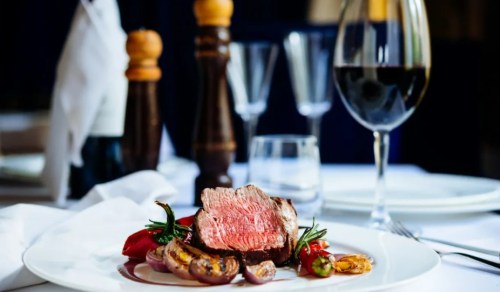 A beautifully plated dish featuring a slice of medium-rare steak, garnished with vegetables and a glass of red wine.