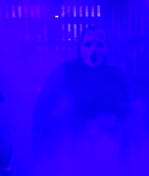 A figure in a mask stands in a foggy, dimly lit space with purple lighting, creating a spooky atmosphere.