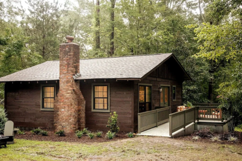 A cozy wooden cabin surrounded by trees, featuring a brick chimney and a small deck.