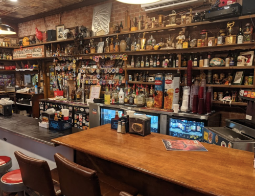 A rustic bar with a wooden counter, shelves filled with bottles, and various memorabilia on the walls.