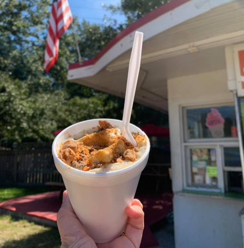 A hand holds a cup of dessert topped with crumbled cookies, with a flag and a food stand in the background.