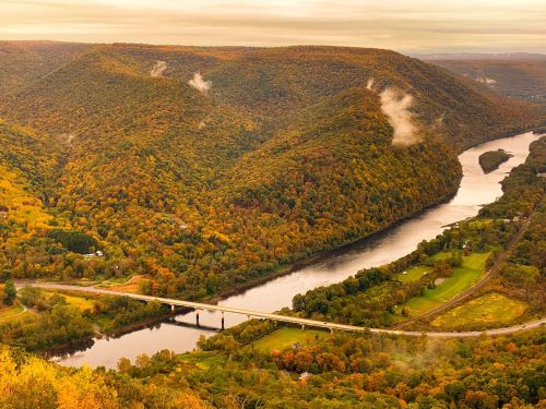 A scenic view of rolling hills with autumn foliage, a winding river, and a bridge under a cloudy sky.