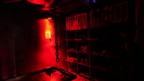 A dimly lit room with red lighting, featuring a warning sign, a chain, and shelves with various items.