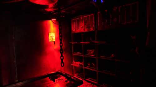 A dimly lit room with red lighting, featuring a warning sign, a chain, and shelves with various items.