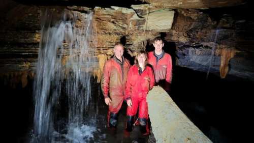 Three people in red caving suits stand near a waterfall in a cave, surrounded by rock formations and water.