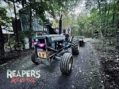 A blue tractor with a trailer parked on a gravel path surrounded by trees, with a spooky logo in the corner.