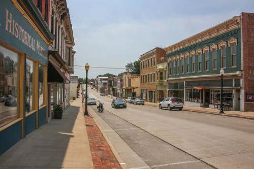 A quiet street lined with historic buildings, shops, and parked cars under a clear blue sky.