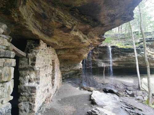 A rocky cave entrance with a stone wall, surrounded by trees and a small waterfall in the background.