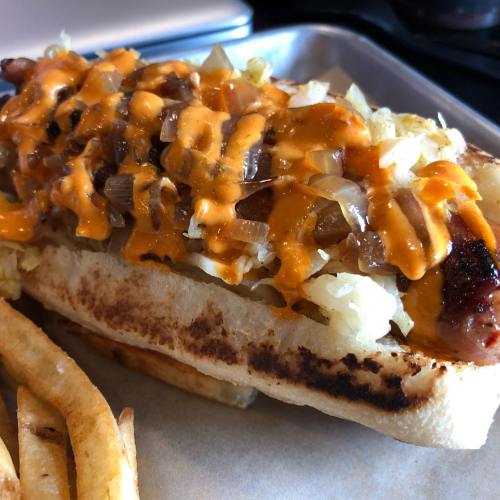 A grilled hot dog topped with onions and yellow sauce, served with a side of golden fries on a metal tray.