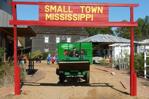 A green tractor drives under a red sign reading "SMALL TOWN MISSISSIPPI" in a rustic, outdoor setting.