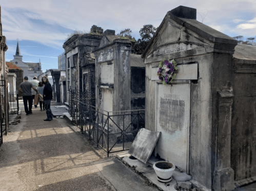 A cemetery path lined with tombs, featuring a flower-adorned grave and visitors walking in the background.