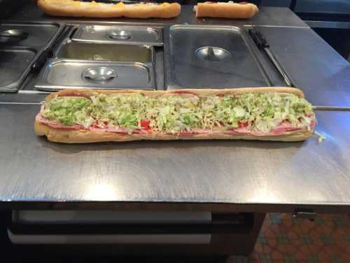 A long sub sandwich filled with layers of lettuce, tomatoes, and deli meats on a stainless steel countertop.