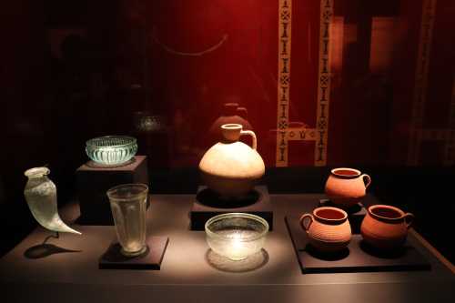 A display of various ancient pottery and glass artifacts on black pedestals against a red background.