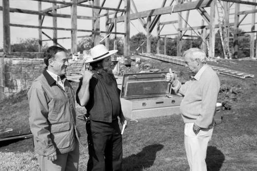 Three men converse at a construction site, with a partially built structure in the background. One man gives a thumbs up.