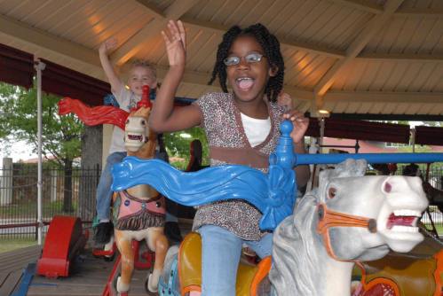 Two children joyfully riding a carousel, waving their hands while sitting on colorful horse figures.
