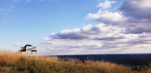 A wooden lookout tower on a grassy hill under a blue sky with fluffy clouds, overlooking a vast landscape.