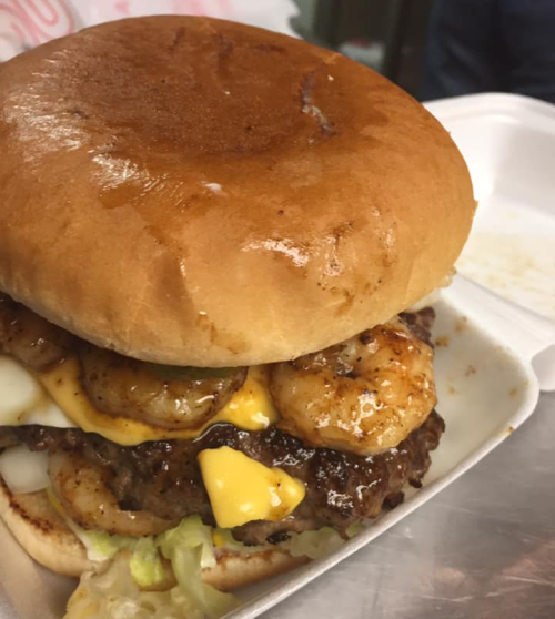 A close-up of a burger with a shiny bun, beef patty, shrimp, cheese, and lettuce in a takeout container.