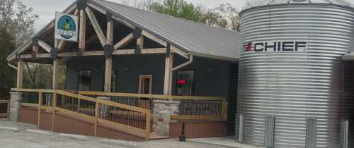 A rustic building with a wooden porch and a large metal grain silo nearby, featuring a sign with a duck logo.