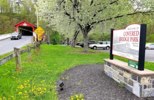 A sign welcomes visitors to Covered Bridge Park, with a red covered bridge and blooming trees in the background.
