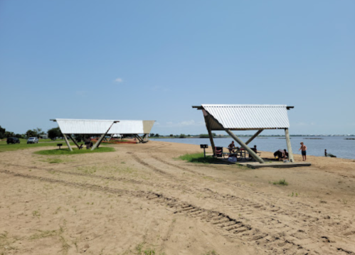 Two modern shelters on a sandy beach by a calm body of water, with a clear blue sky in the background.